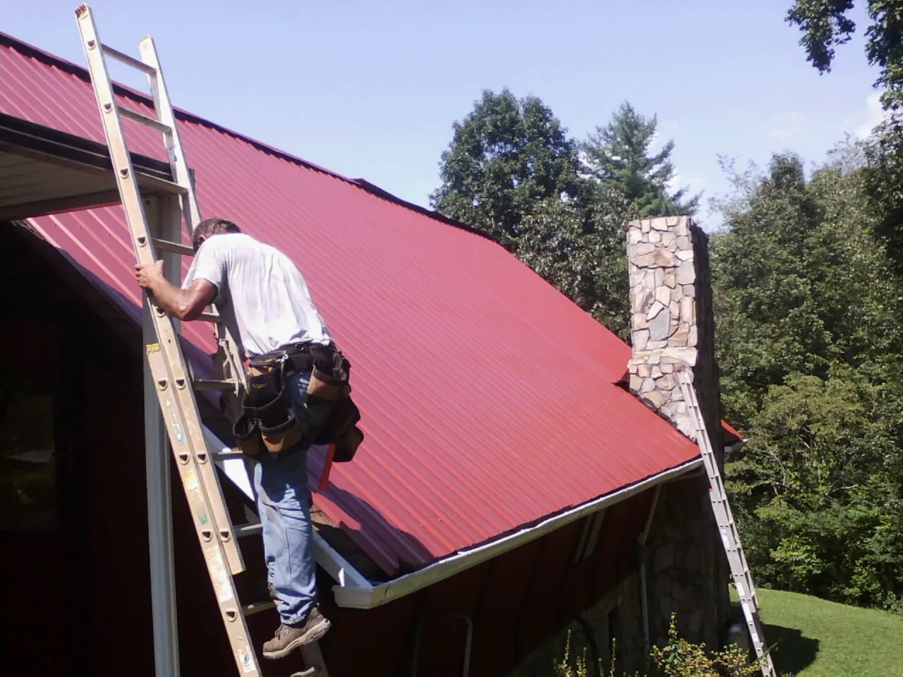 Roofer on a ladder beside a red metal roof and stone chimney — Soffit & Fascia Repair by Roofing Brothers in Bala Cynwyd